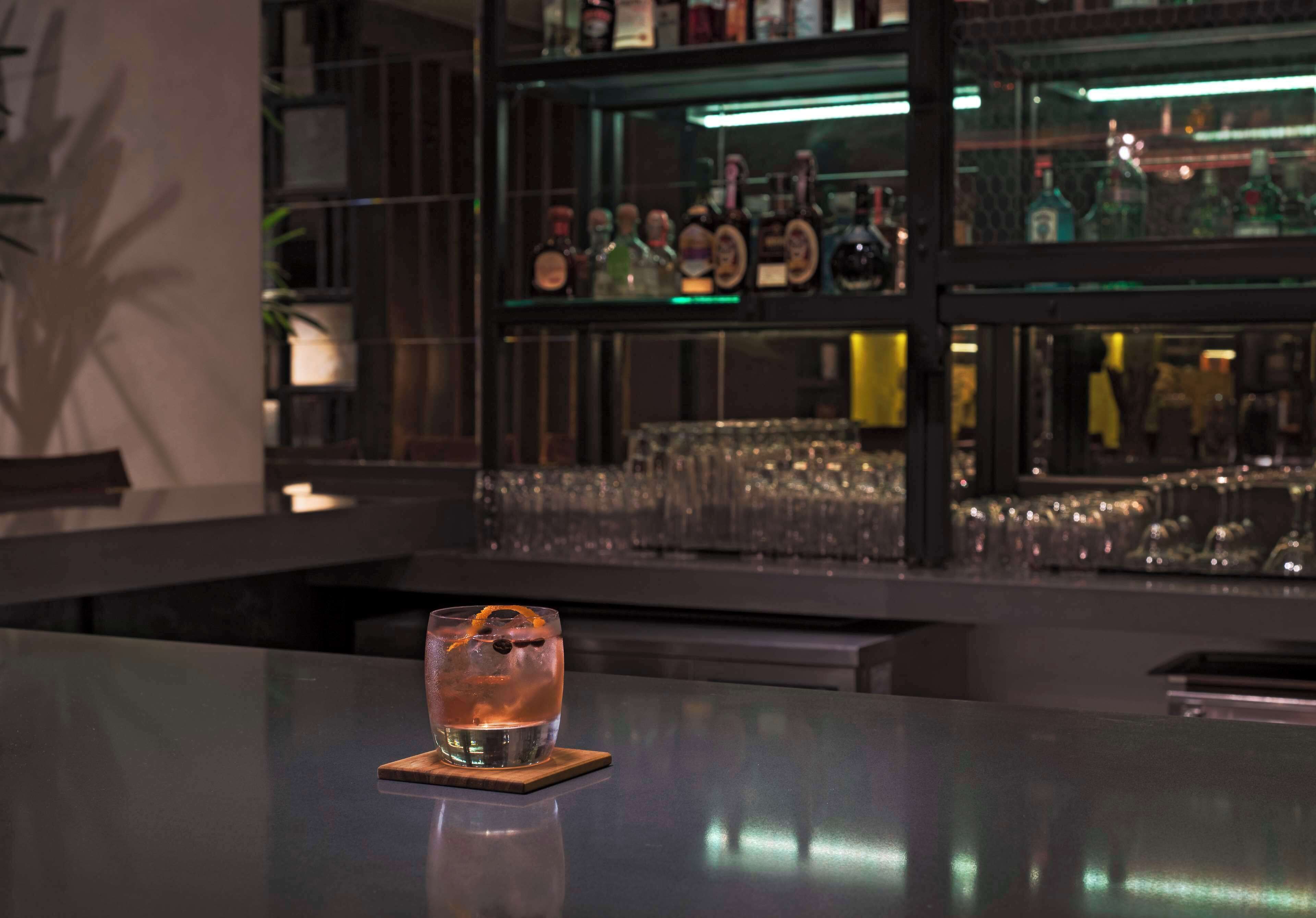 A bar area with a glass of drink on a coaster, shelves with bottles, and glasses in the background.