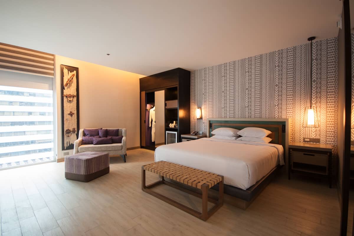 Une chambre d’hôtel moderne dotée d’un grand lit, d’un coin salon avec un canapé et un pouf, d’une armoire et offrant une vue sur la ville par la fenêtre.