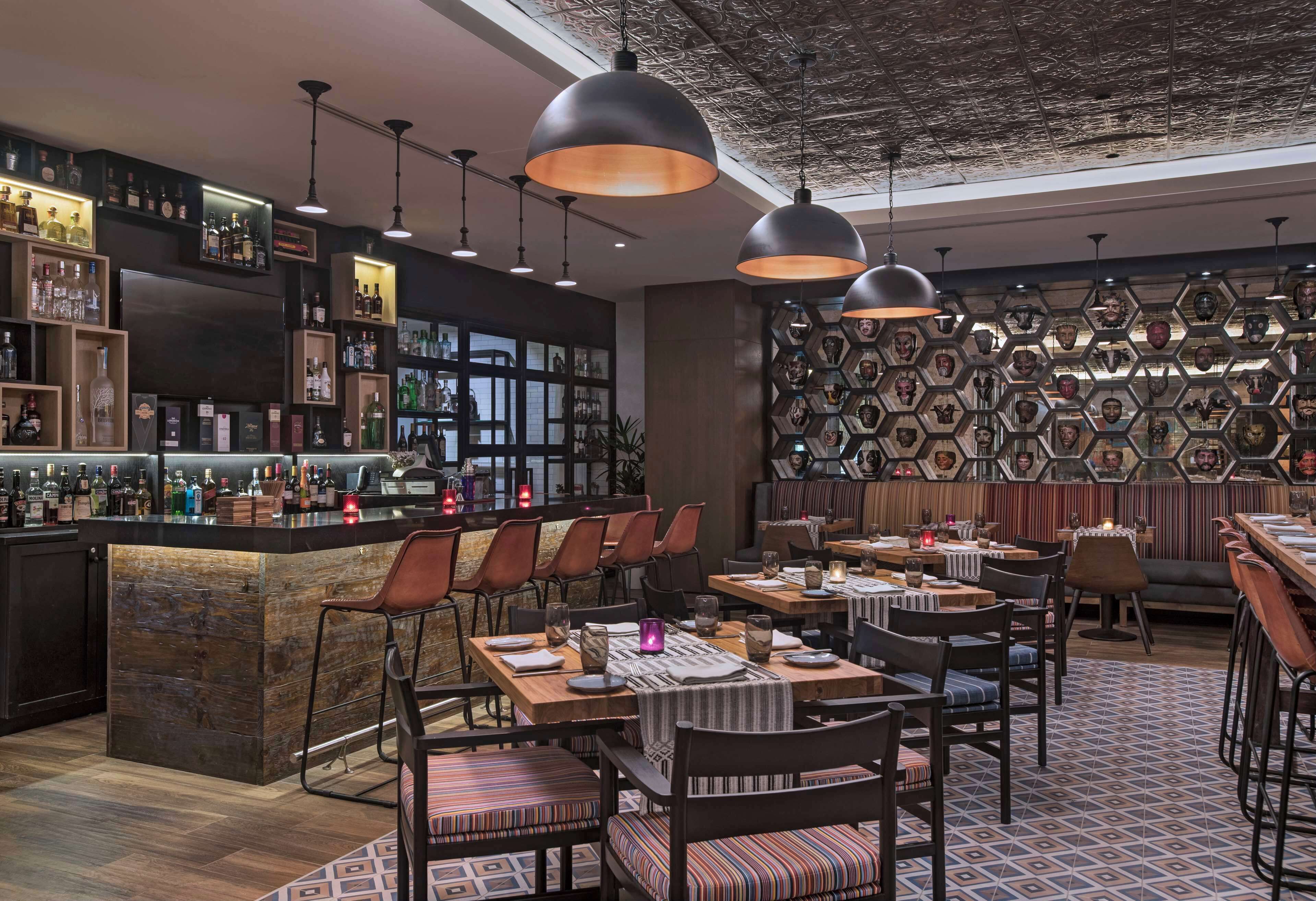 Zona de bar moderna con mostrador de madera, diversas opciones de asiento y una pared decorativa con patrones hexagonales.