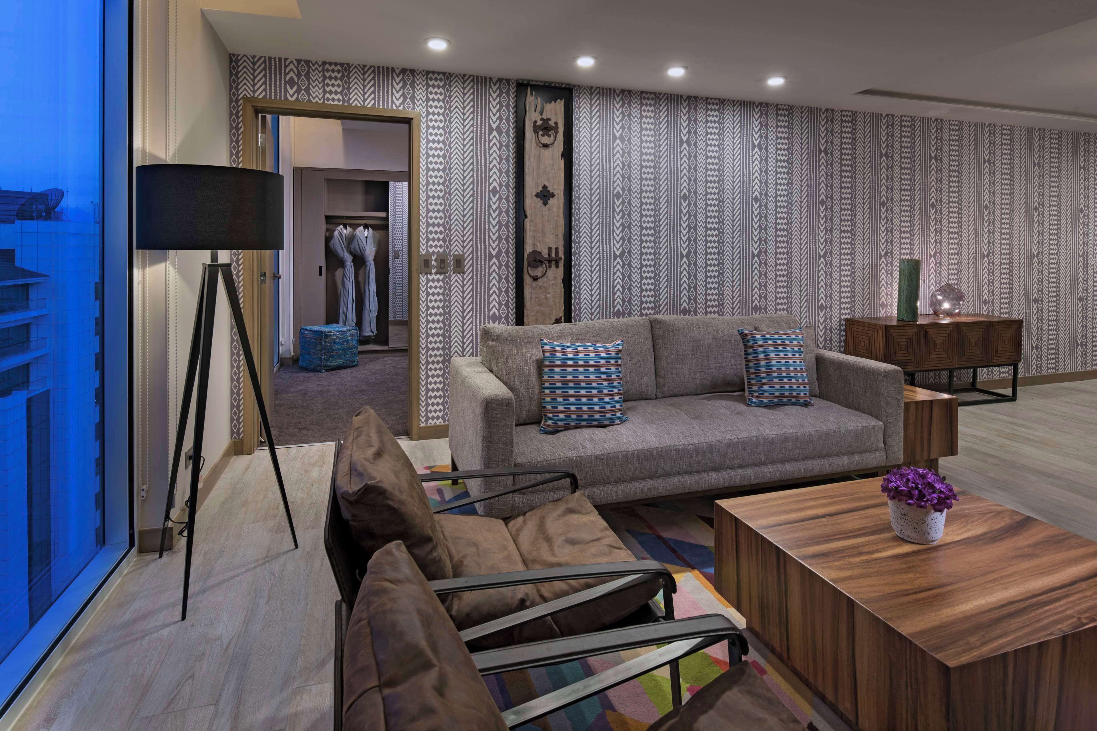 A modern living room with a sofa, armchairs, a coffee table, and a floor lamp. There is a patterned wallpaper and a wardrobe in the background.