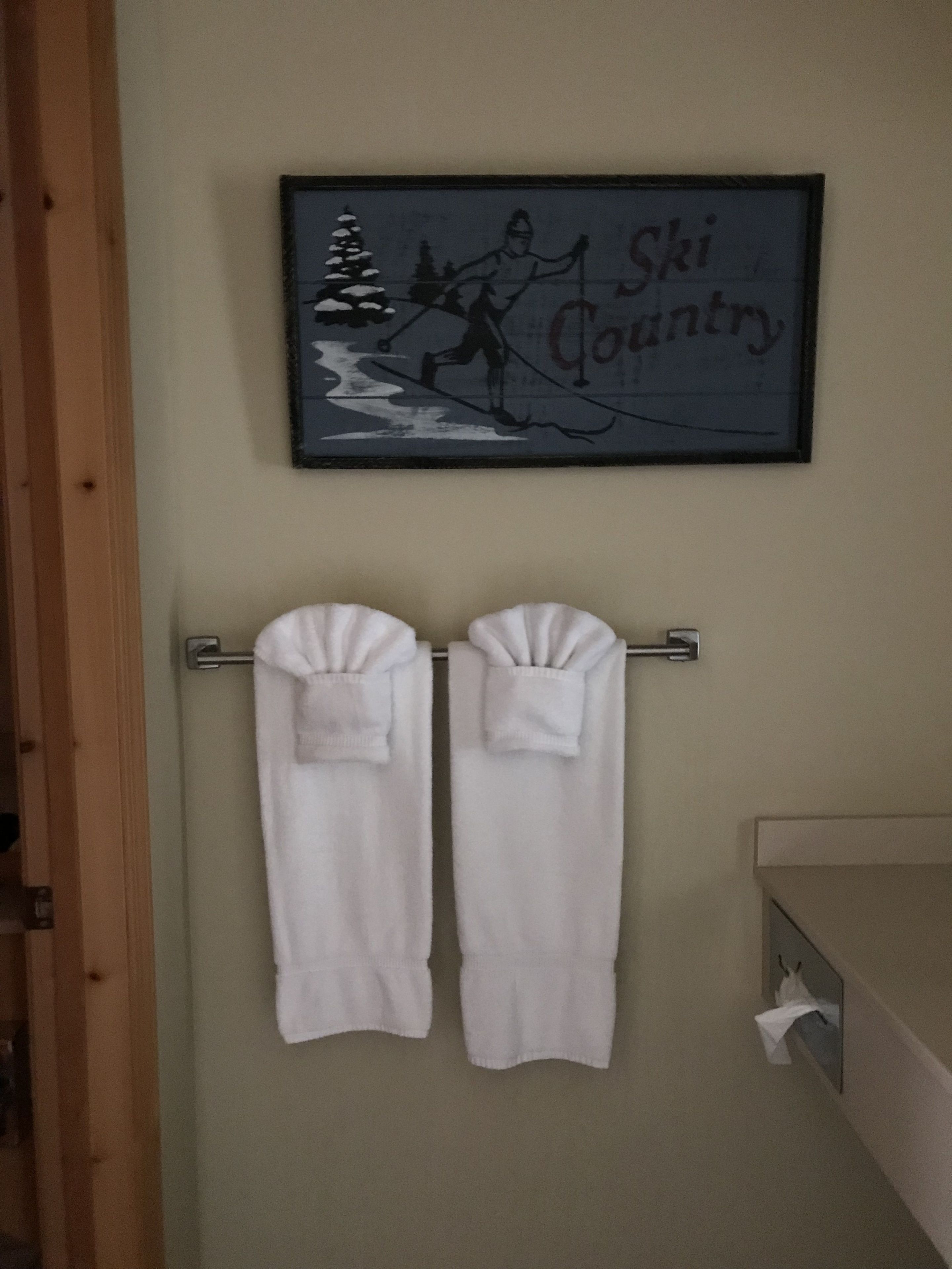 Une salle de bain avec deux serviettes blanches suspendues à un porte-serviettes en métal, un tableau encadré représentant un skieur sur une montagne et un cadre de porte en bois.