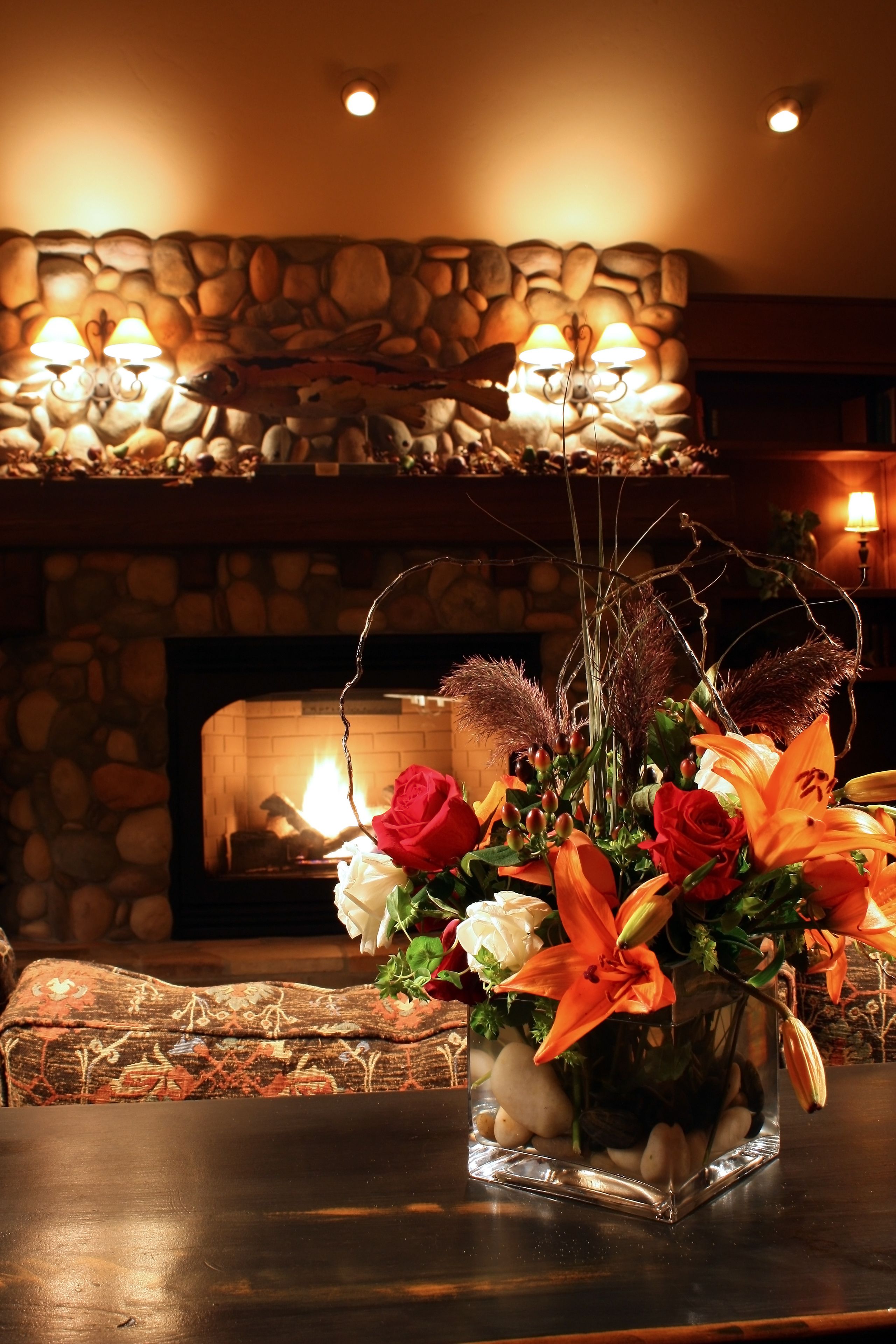 A vase with flowers on a table in a warmly lit room with a stone wall and fireplace.