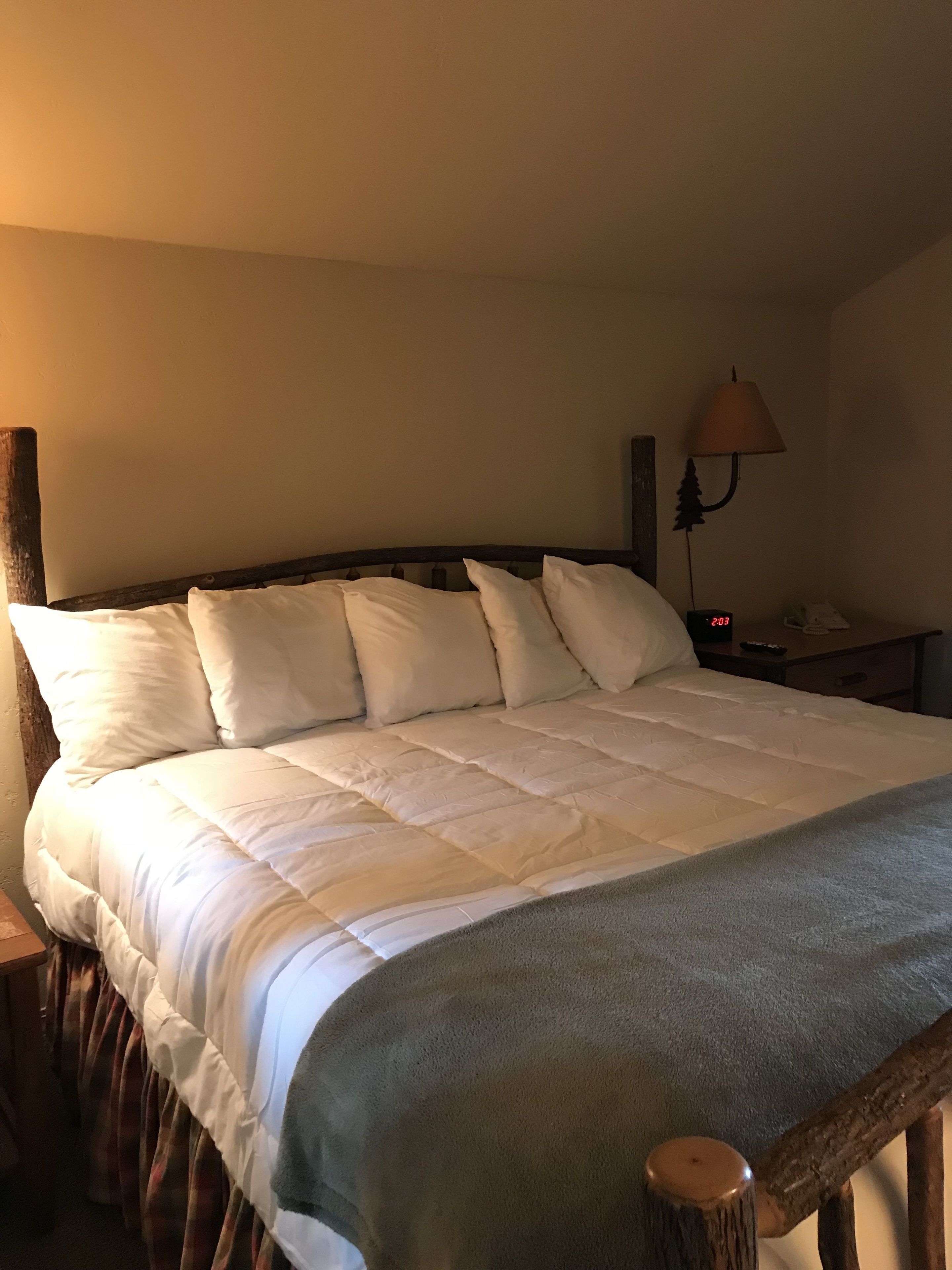 A neatly made bed with a wooden headboard, a bedside lamp, and a digital clock displaying 2:03.