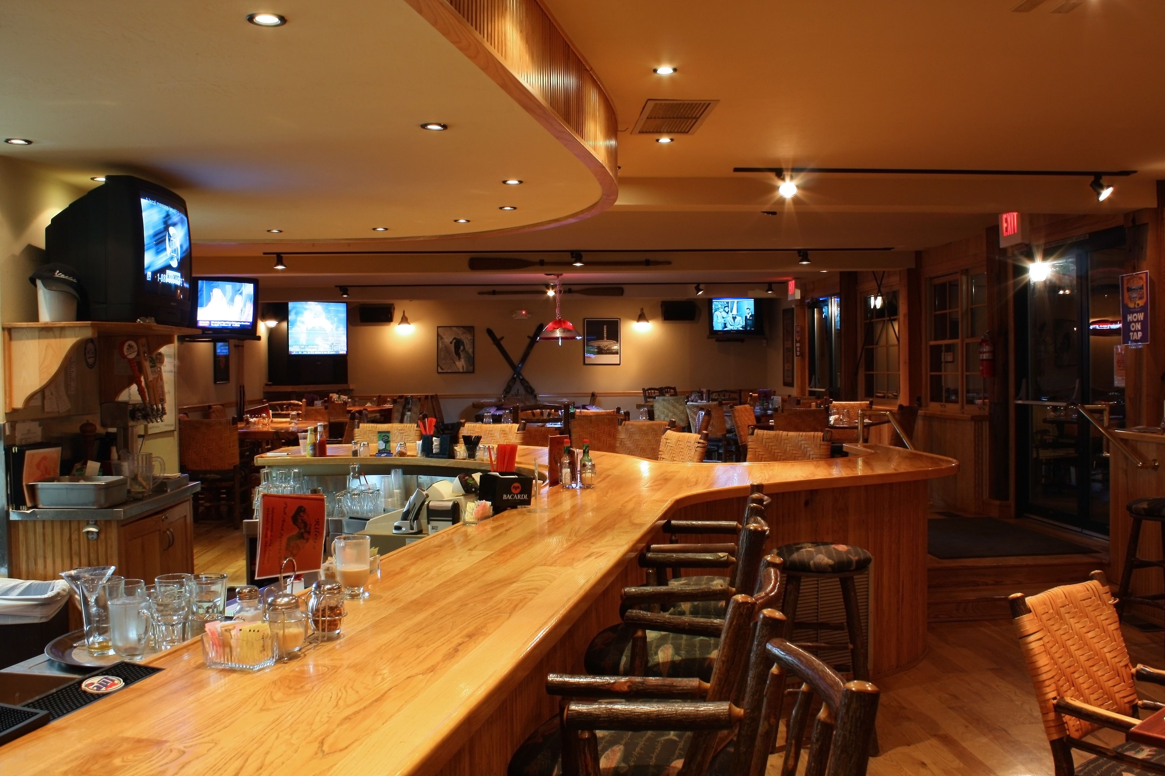 A bar area with wooden bar counter and stools, multiple televisions mounted on the wall, and a variety of bar tools and glasses on the counter.