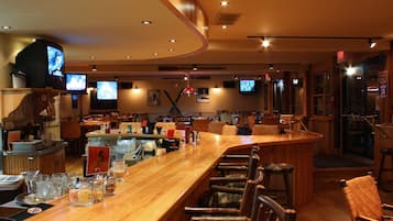A bar area with wooden bar counter and stools, multiple televisions mounted on the wall, and a variety of bar tools and glasses on the counter.