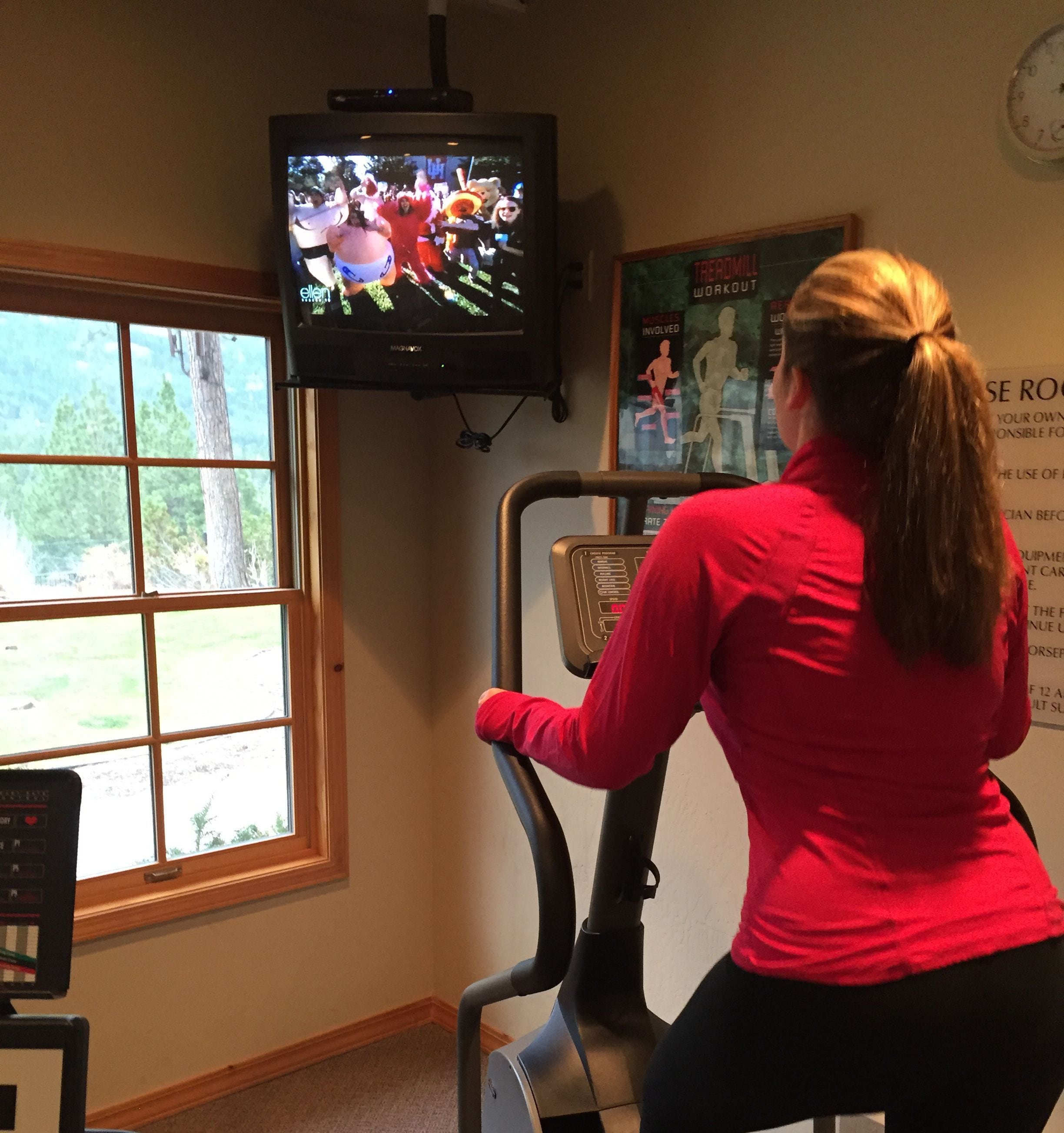 A woman using an exercise bike in a room with a window, a treadmill, and a poster on the wall.