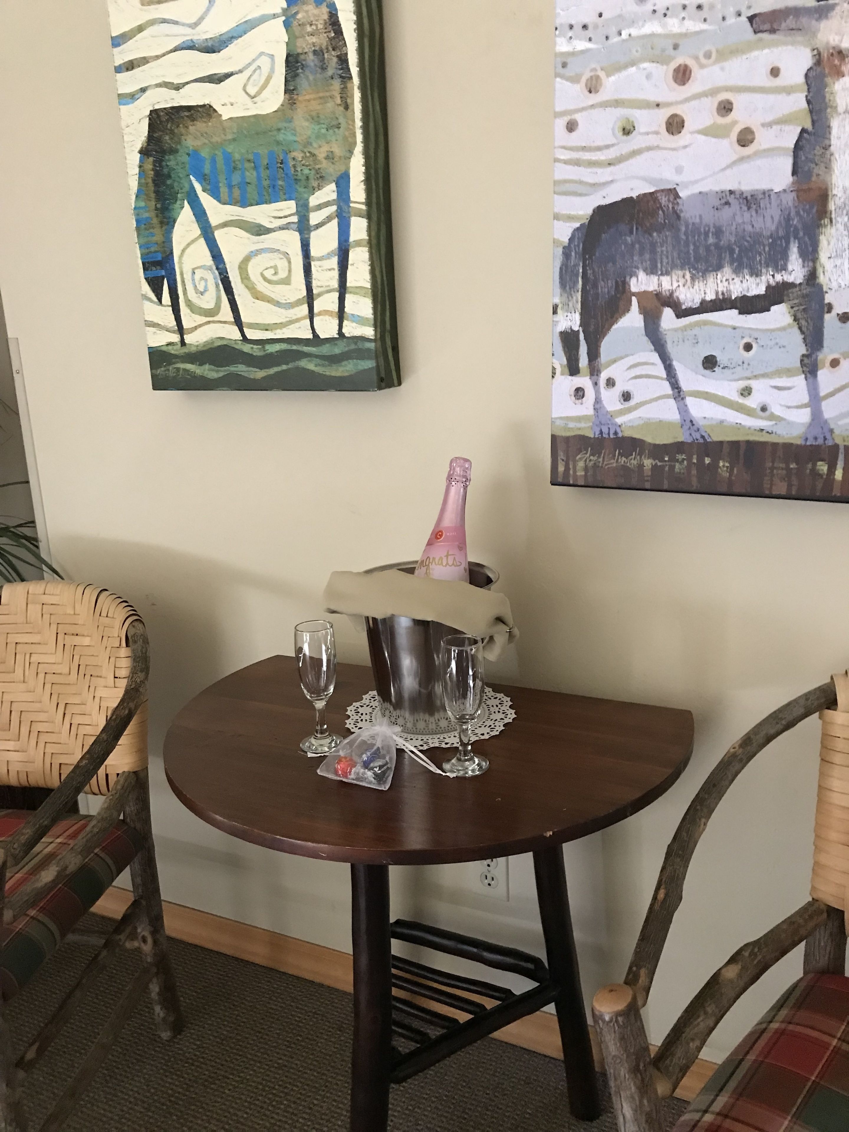 A wooden table with a metal bucket, two wine glasses, and a plastic bag with a red item.