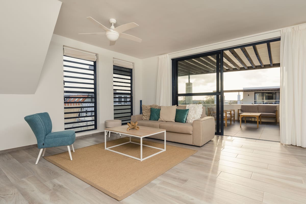 Ein modernes Wohnzimmer mit einer Couch, einem Couchtisch und einem Sessel. Es gibt ein großes Fenster mit Jalousien, einen Deckenventilator und einen Blick ins Freie.