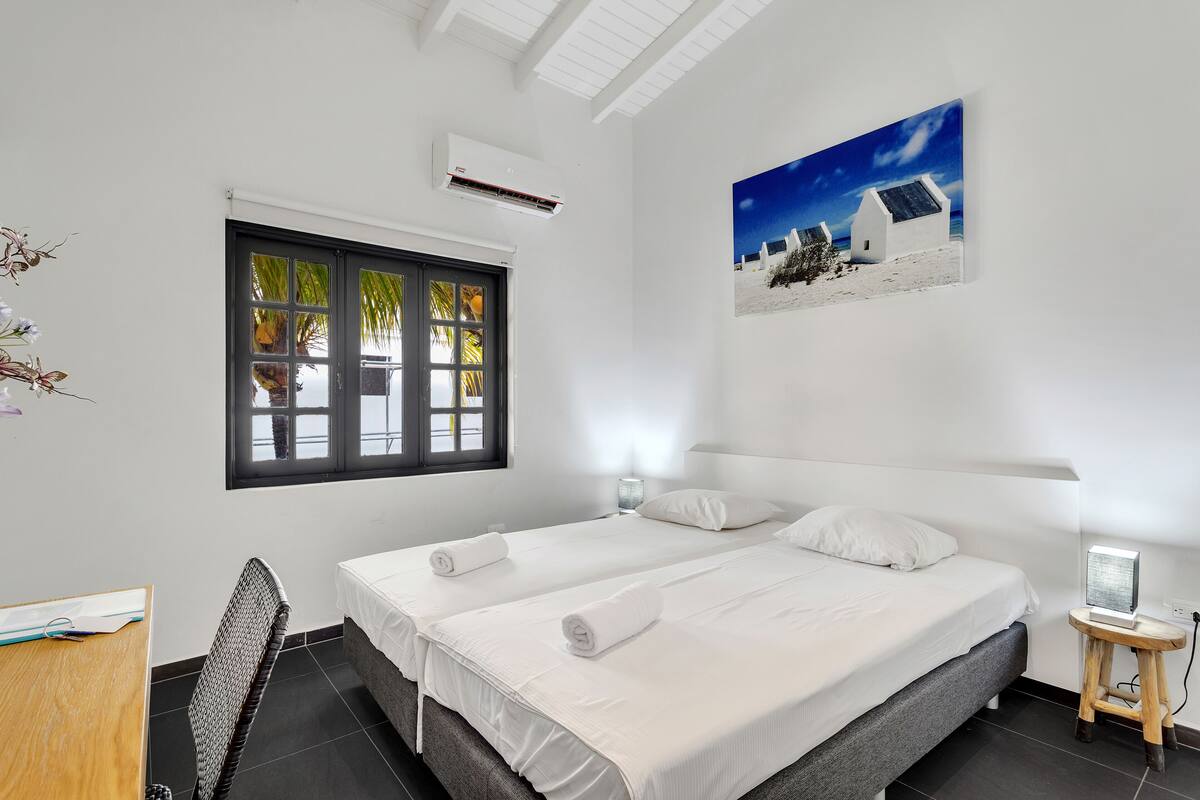 Ein Hotelzimmer mit zwei Betten, einem Schreibtisch, einem Stuhl, einem Fenster mit Blick auf Palmen und einem Bild von einer Strandlandschaft.