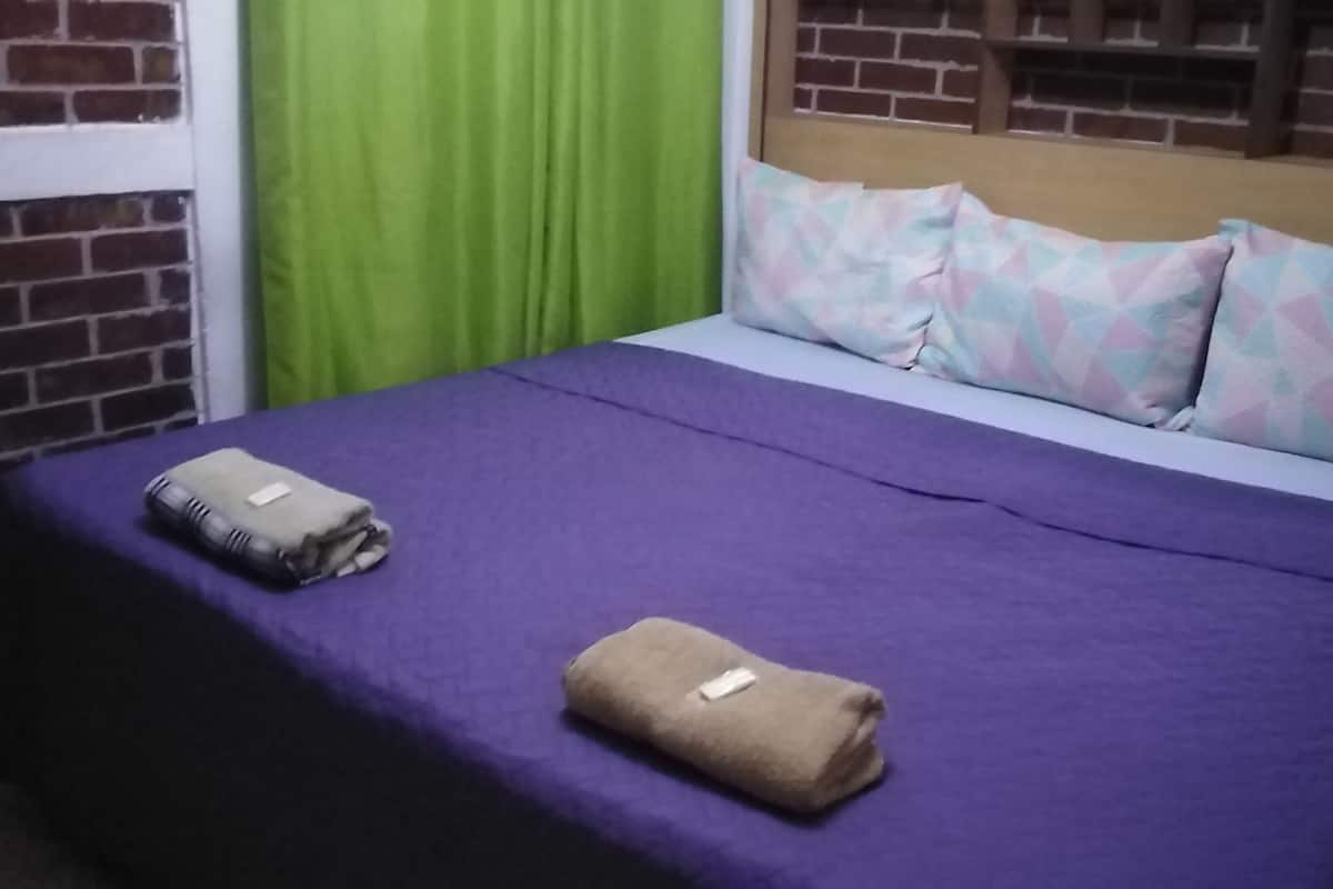 A bed with a purple bedspread, two folded towels, and patterned pillows.
