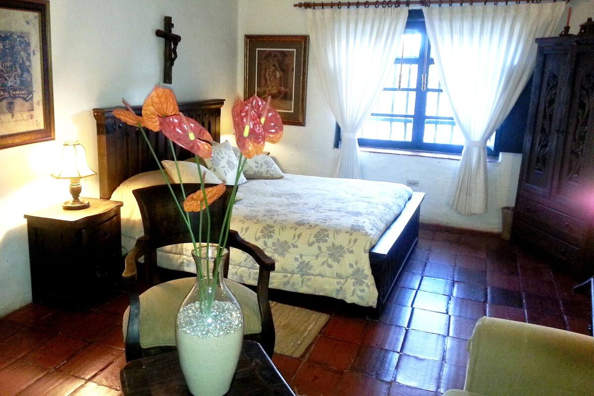 Un dormitorio con una cama, una silla, una mesa con un florero, una lámpara y una ventana con cortinas.