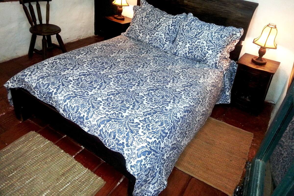 Un dormitorio con una colcha estampada, dos lámparas de noche, una silla de madera y un tapete tejido.