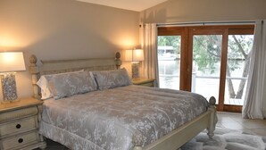 A bedroom with a large bed, two bedside tables with lamps, a view of a body of water through a glass door, and a patterned rug. - New Orleans Area Waterfront Retreat 117 Near the French Quarter/ Bourbon Street (Slidell)