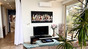 A modern living room with a flat-screen TV, a large framed cityscape picture, a small table with a vase of flowers, and a large window with a view of greenery. - Signature 406 Private Pool Sept 18-23 @ -50%, Oct 18-23 @ -40%, Nov 18-22 @ -25% (Puerto Vallarta,)