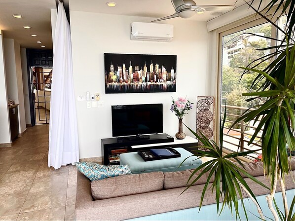 A modern living room with a flat-screen TV, a large framed cityscape picture, a small table with a vase of flowers, and a large window with a view of greenery.