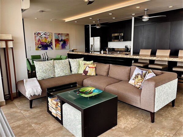 A modern living room with a sectional sofa, a coffee table, and a kitchen area in the background.