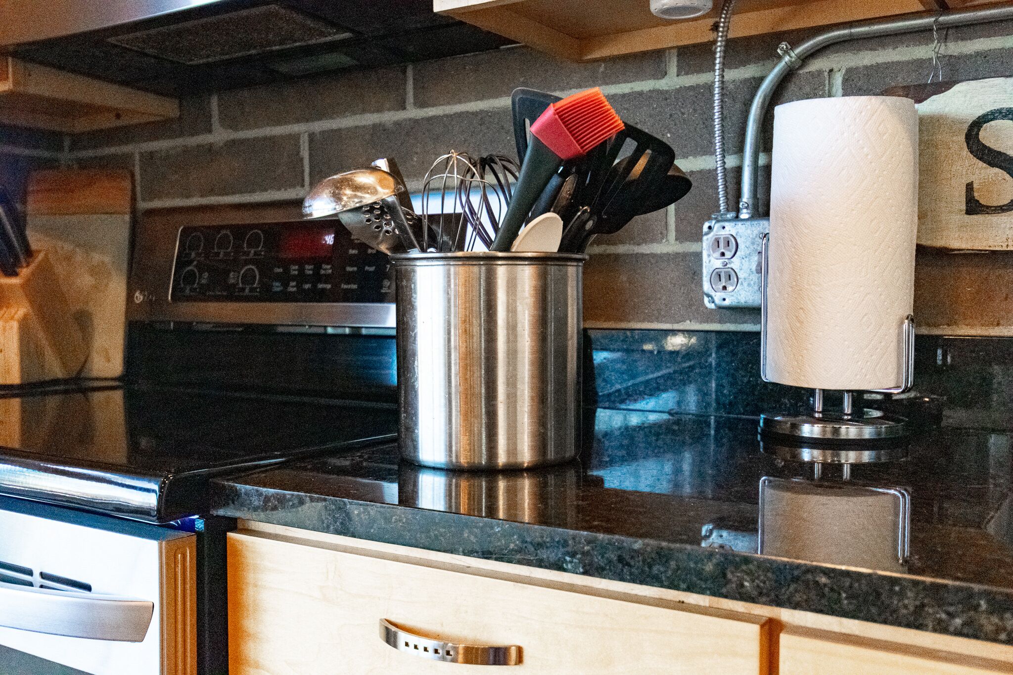A kitchen with a stainless steel utensil holder on the counter, a paper towel dispenser, and a brick wall backsplash.