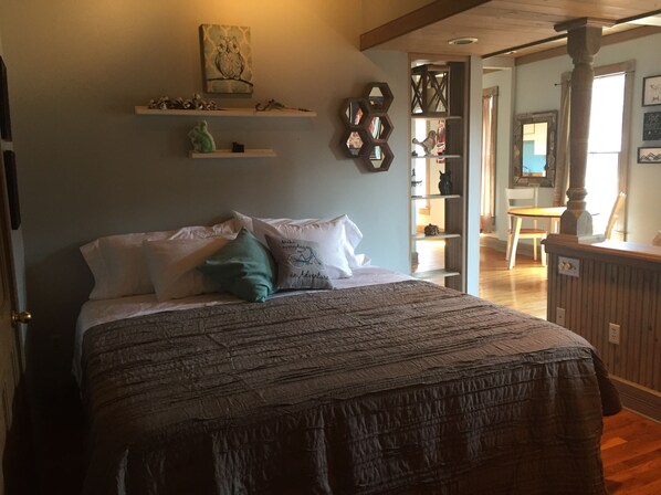 A bedroom with a bed, pillows, a shelf with decorative items, and a view into another room.