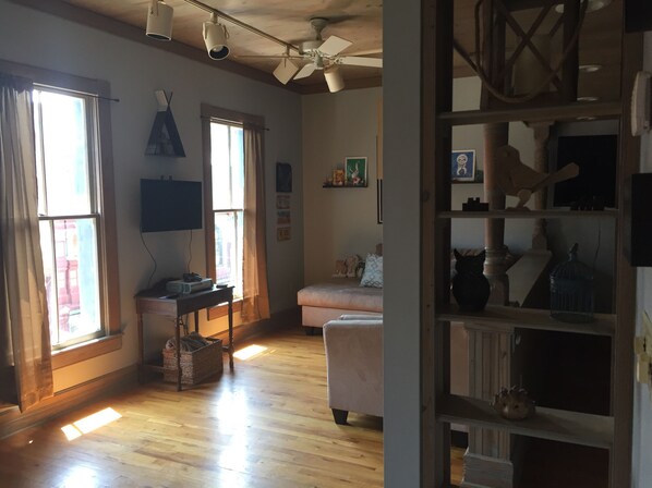 Una habitación amplia con suelo de madera, ventilador de techo y ventanas grandes con cortinas. Hay un escritorio con una computadora y una cesta en el suelo.