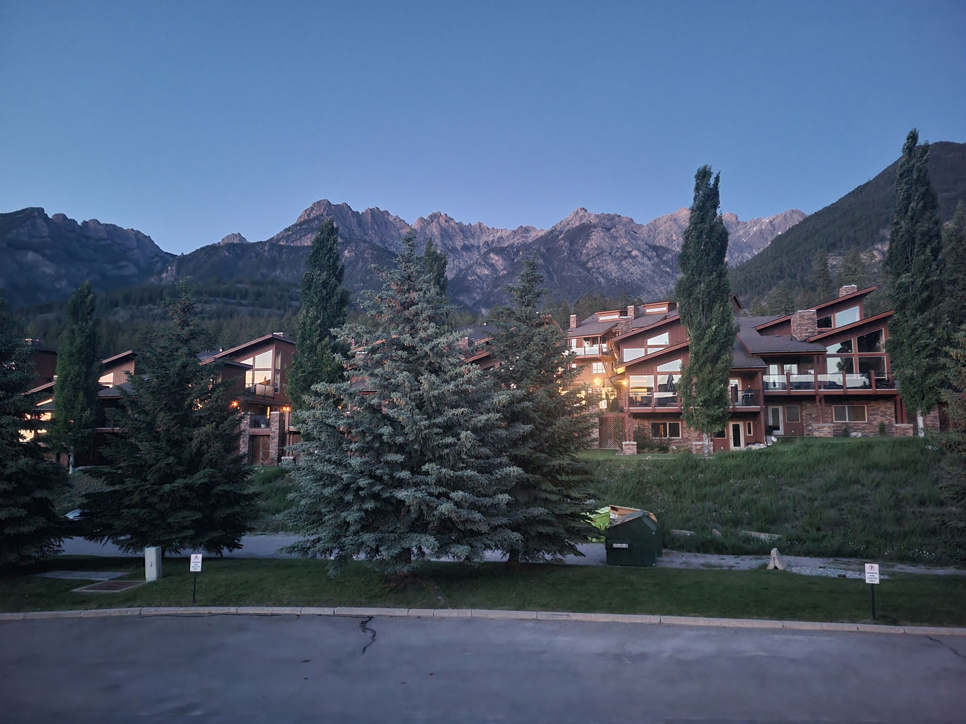 A multi-story lodge with a mountain backdrop, featuring large windows and a prominent evergreen tree in the foreground.
