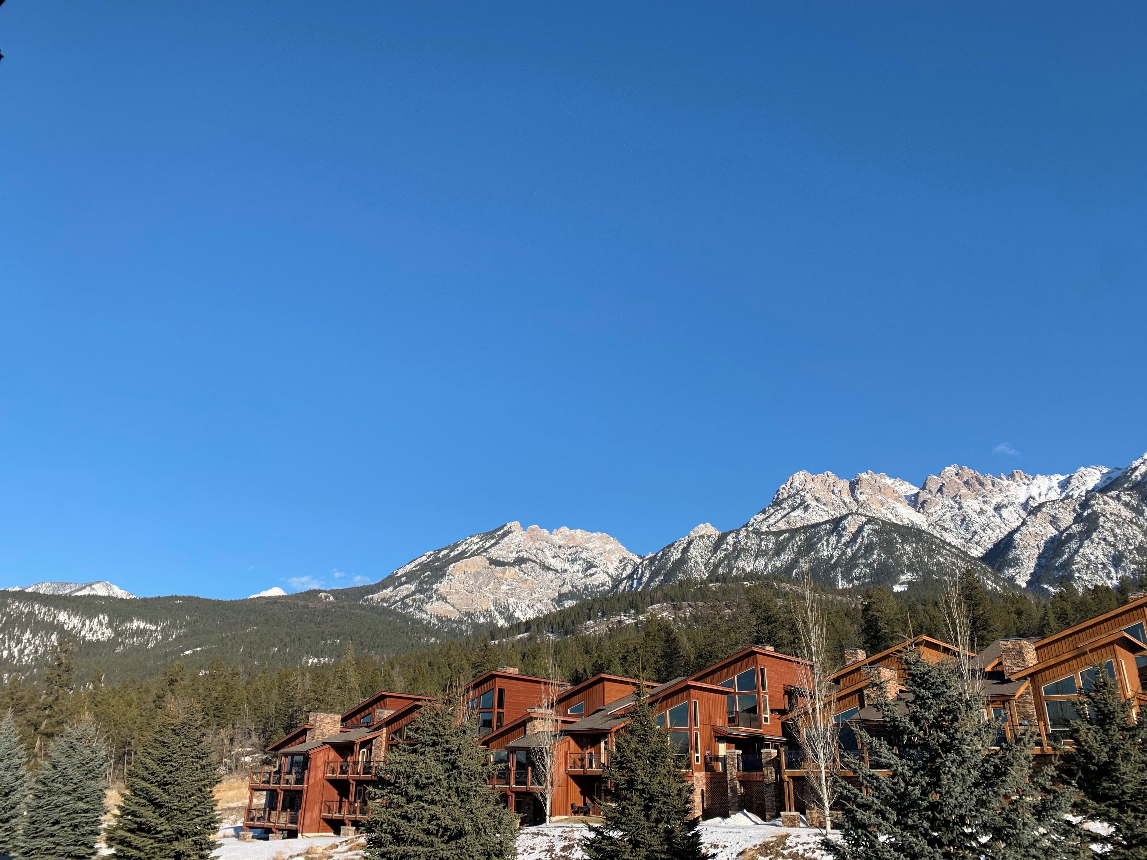A mountain lodge with multiple wooden buildings, surrounded by snow-covered mountains and evergreen trees.