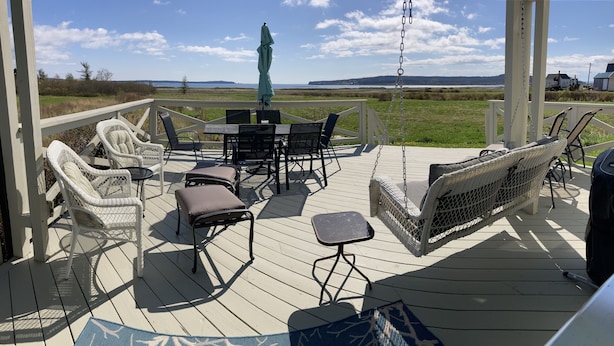 A wooden deck with wicker furniture, including chairs and a swing, overlooking a grassy area and water. - Classic Maine Oceanfront Cottage, Gorgeous Views, Centrally Located, Pets, A/C (Lubec)