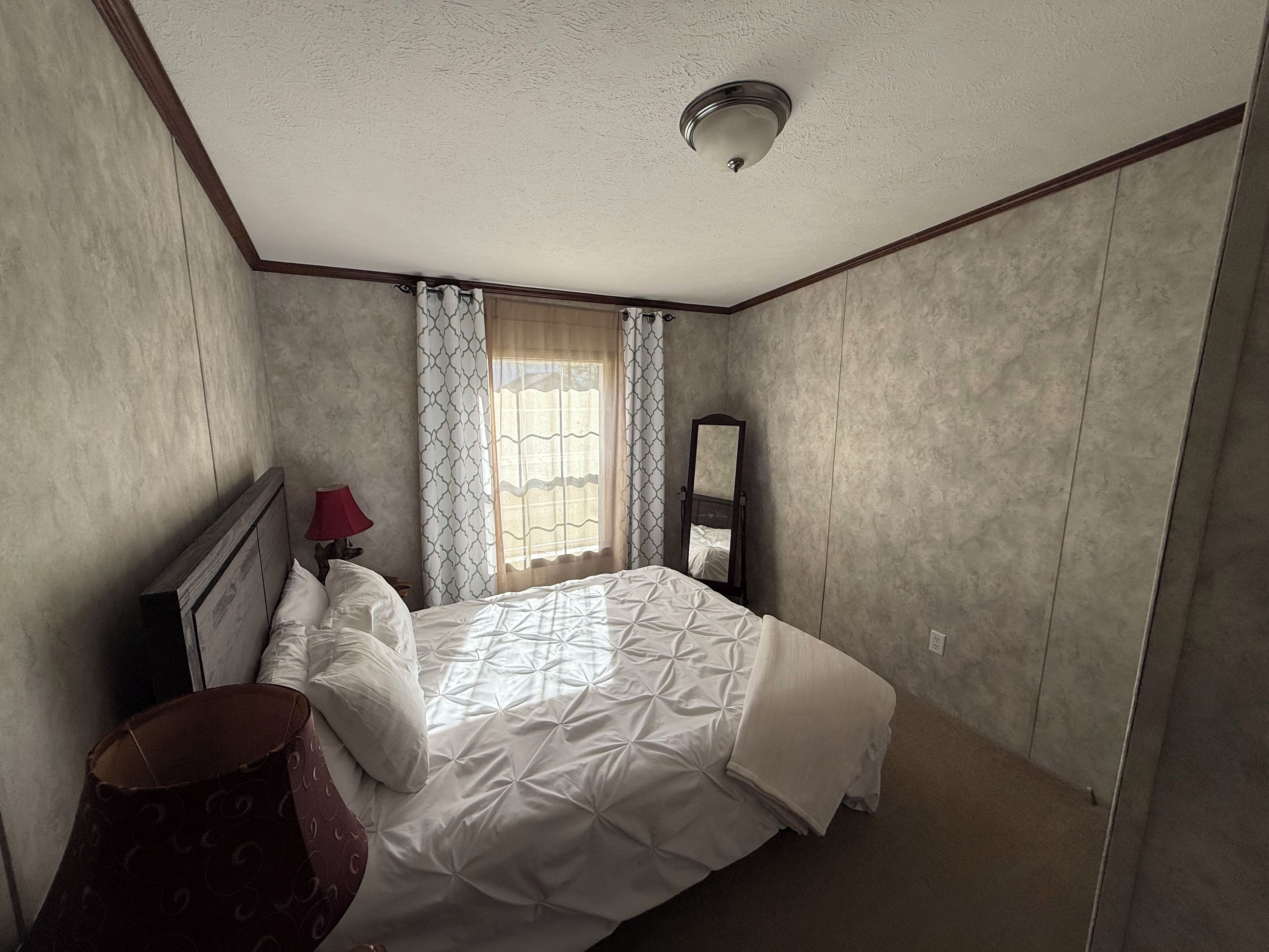 A bedroom with a bed, a nightstand, a lamp, a mirror, and a window with curtains.