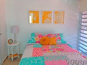 Ein Schlafzimmer mit einer farbenfrohen Stegdecke, einem Nachttisch mit Lampe und drei gerahmten floralen Kunstwerken an der Wand.