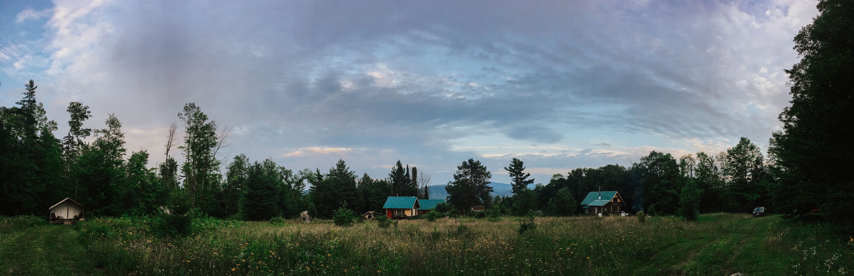 Maaseutumaisema, jossa on useita mökkejä, ja niitä ympäröi tiheä metsä. Taivas on pilvinen.
