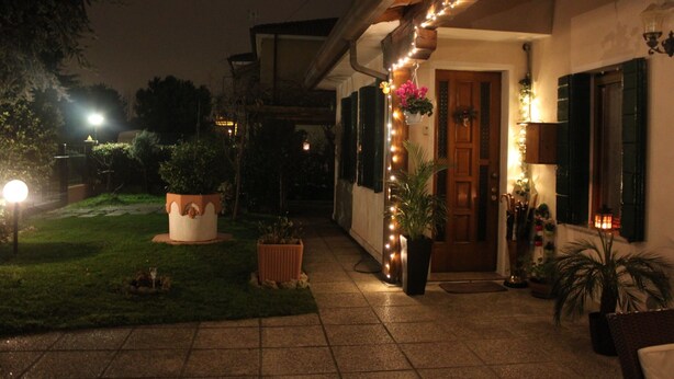 A nighttime view of a building's exterior with a well-lit entrance, potted plants, and string lights. - Venice Holiday Home, free wi-fi, private garden, romantic and affordable. (Mogliano Veneto)