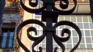 Ein schmiedeeisernes Tor mit aufwendigen Rankenornamenten, vor einem Gebäude mit großen Fenstern und einem blauen Oberteil.