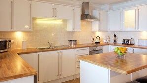 Une cuisine moderne avec des meubles blancs, un plan de travail en bois, un four encastré, une bouilloire et un panier à fruits.