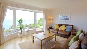 Un salon doté d’une grande fenêtre donnant sur la mer, d’un canapé en cuir marron agrémenté de coussins, d’une table basse blanche et d’une plante en pot.