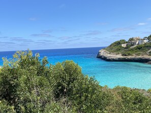 A coastal landscape with clear blue sea, rocky cliffs, and a building with a pathway leading up to it.