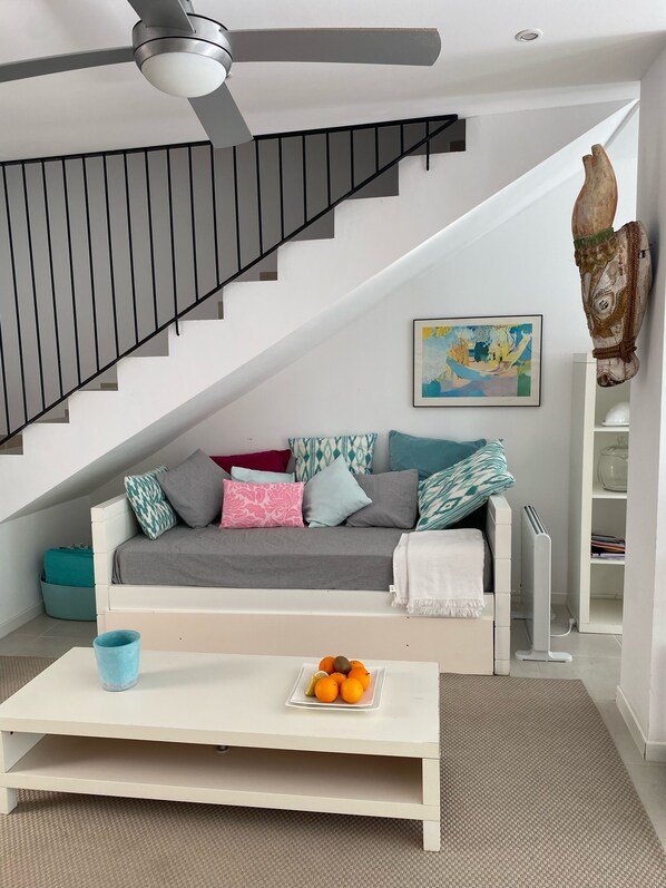 A modern living room with a white sofa, a coffee table with fruit, a black staircase, and a ceiling fan.