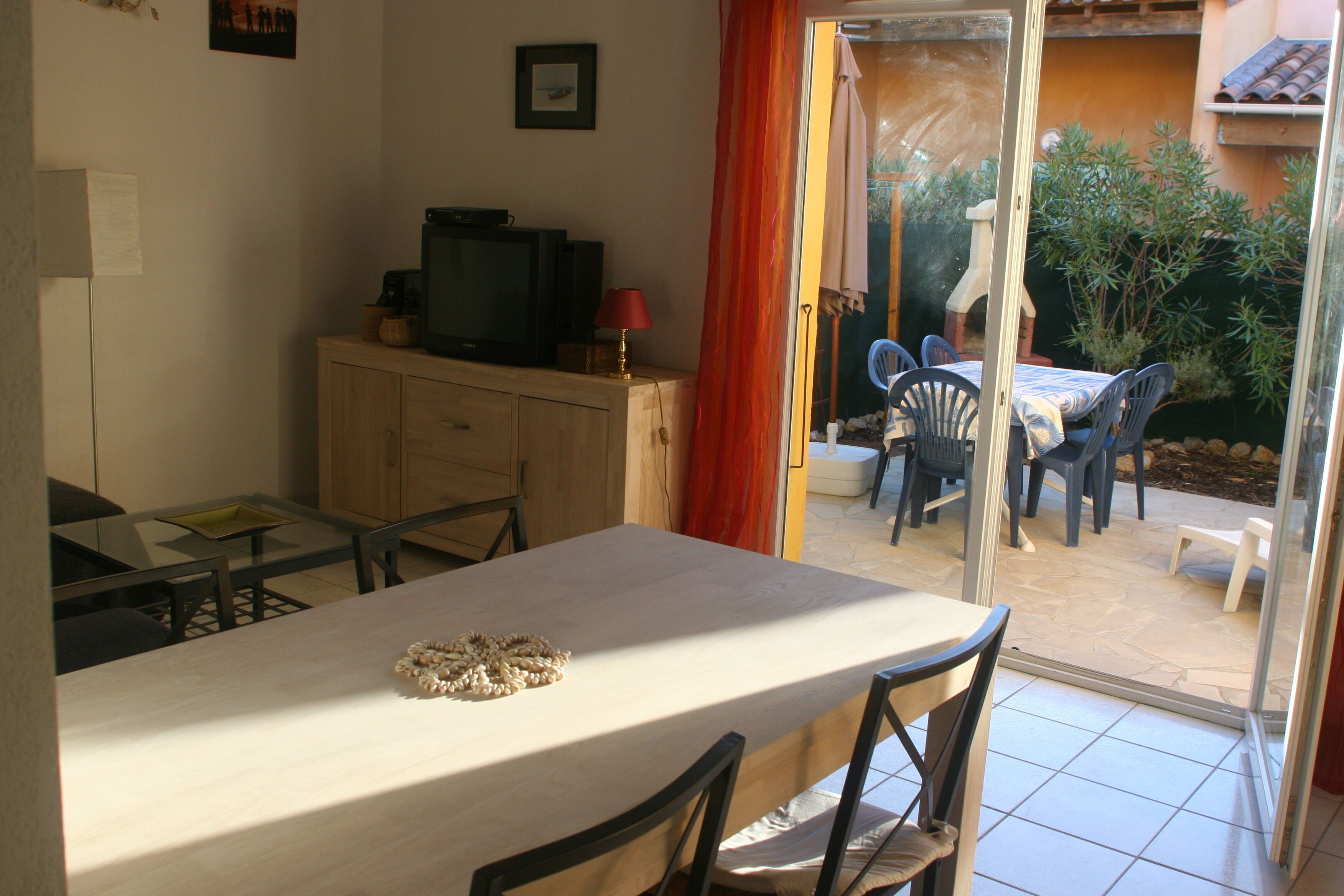 A dining area with a table and chairs, a television on a cabinet, and a view of an outdoor patio with chairs and a table.