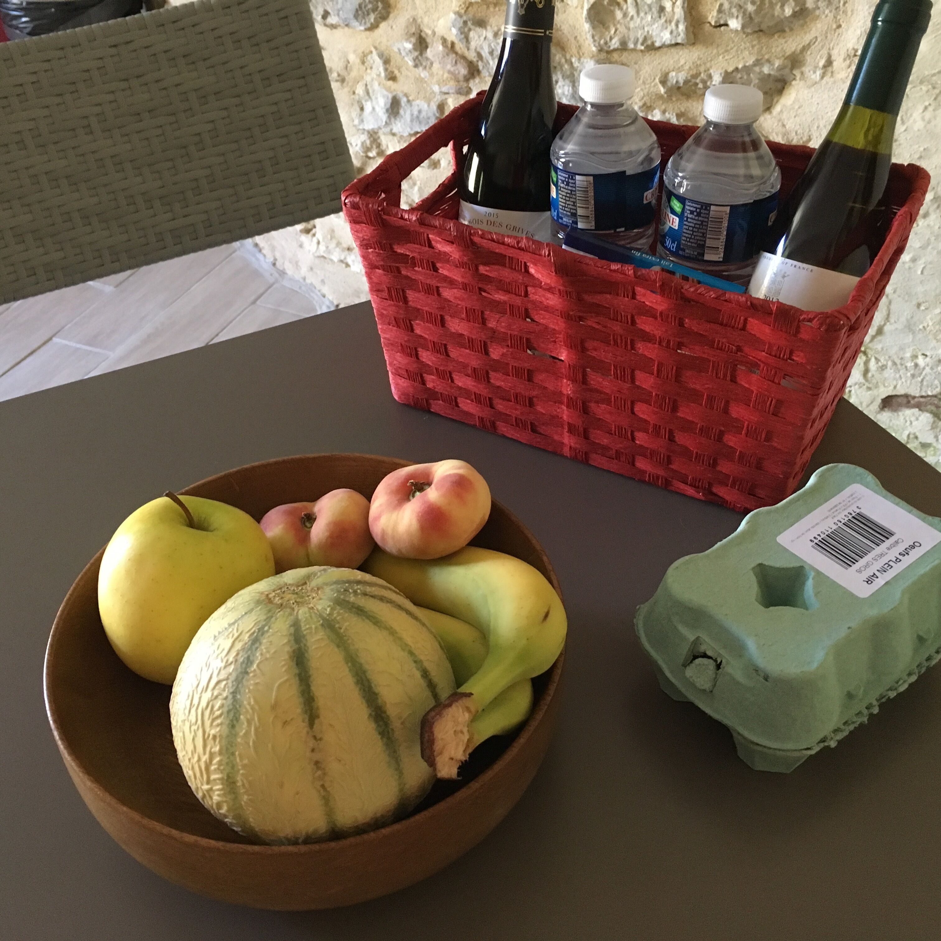 A table with a bowl of fruit, a carton of eggs, and a basket containing bottles and a water bottle.