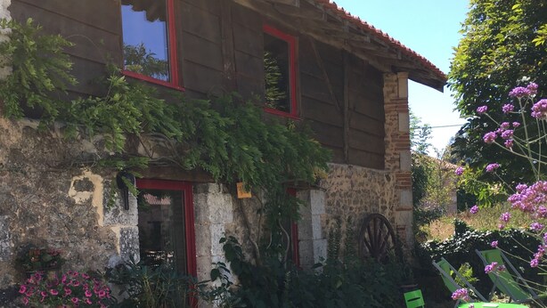 Un edificio de piedra rústica con puerta roja y ventanas, rodeado de follaje y un camino de grava.