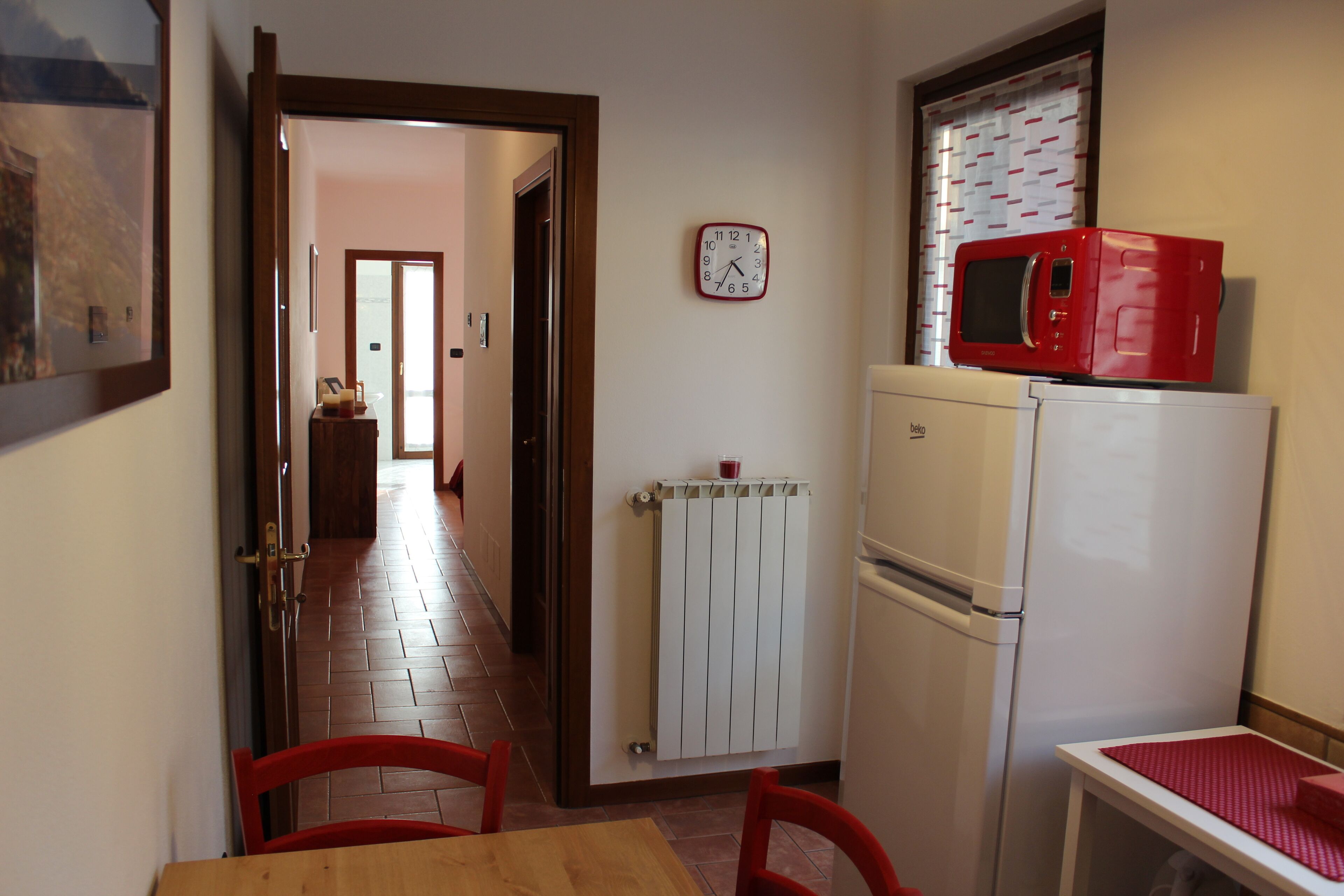 A narrow kitchen with a red microwave, a white refrigerator, a clock, and a door leading to another room.