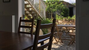 Un espace repas avec une table et des chaises en bois, un escalier menant à un niveau supérieur, et une vue sur un jardin et un bâtiment à l’extérieur.