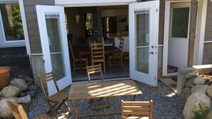 Une terrasse ouverte avec une table et des chaises en bois, une cuisine visible à travers la porte ouverte et un sol recouvert de gravier.