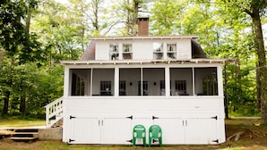 A white wooden cabin with a balcony, a porch, and two green chairs outside.