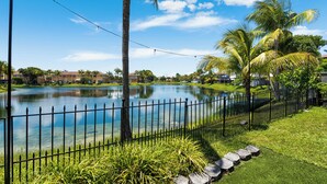 A serene lakeside view with a clear sky, a fence, and palm trees.