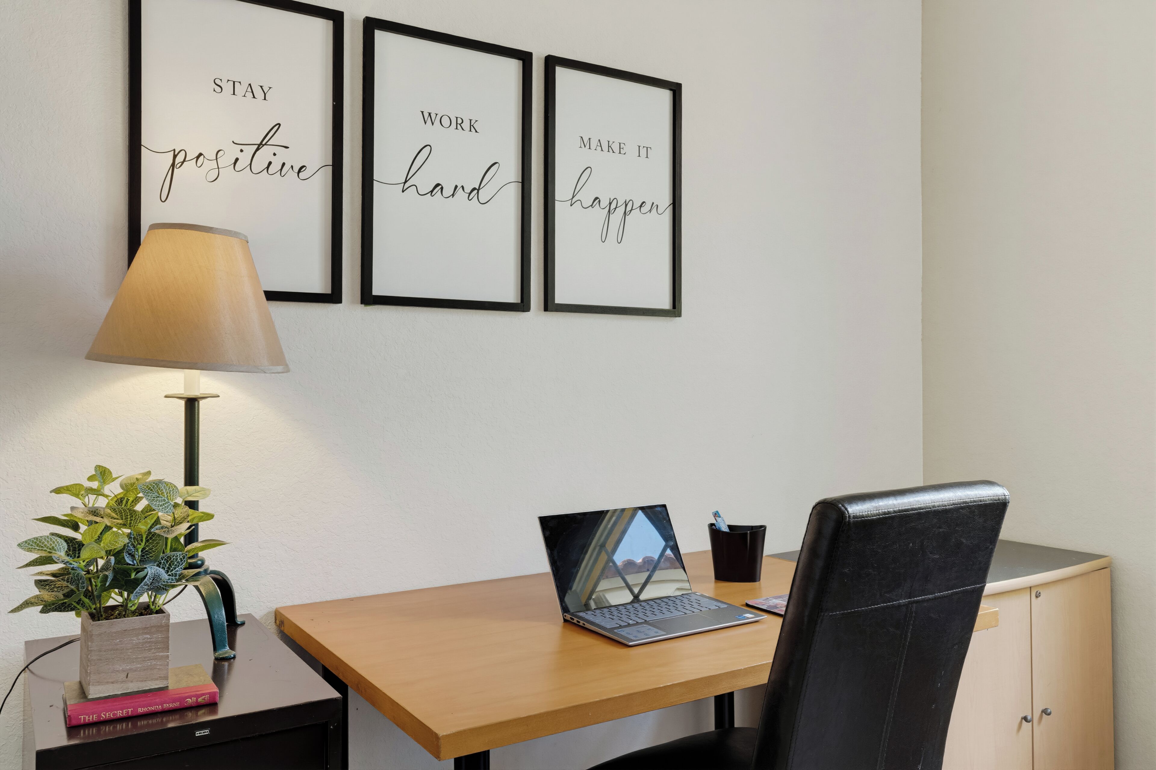 Un espacio de trabajo con escritorio, silla, ordenador portátil, lámpara, citas motivacionales enmarcadas y una planta en maceta.