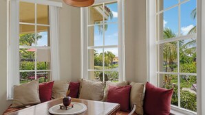 A dining area with a wooden table, cushioned seating, and large windows offering a view of a garden and palm trees.