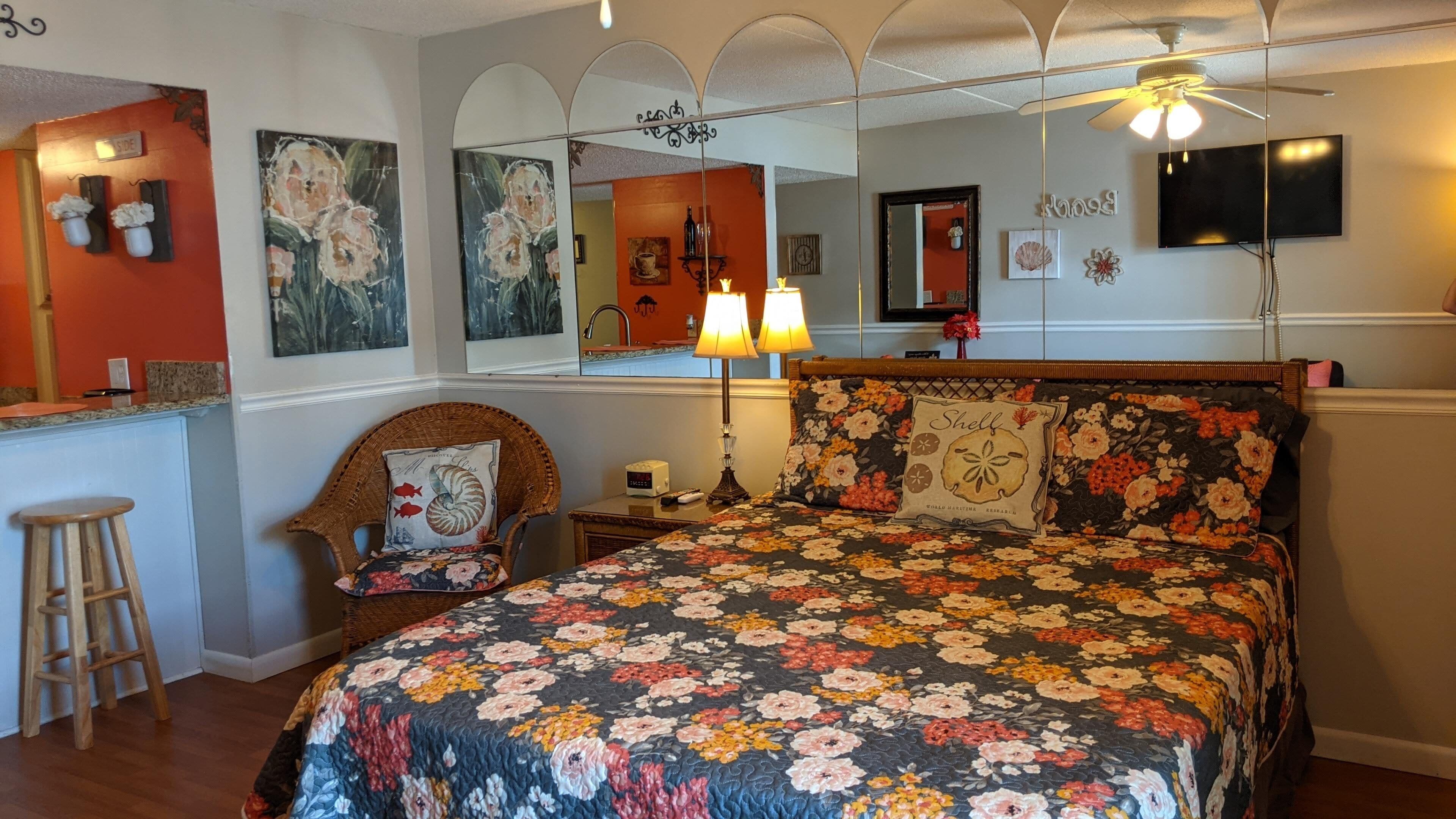 Une chambre à coucher avec une couvre-lit à motifs floraux, une chaise en osier, une table de chevet avec une lampe et un grand miroir au-dessus du lit.