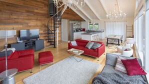 A spacious living room with a red sofa, a grey sofa, and a white armchair. There is a TV on a stand, a chandelier, and a staircase leading to an upper level.