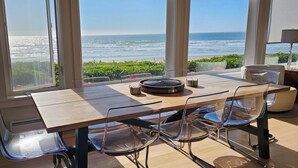 A dining area with a view of the ocean, a wooden table, clear chairs, and a small side table with a lamp.