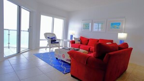 Un salon avec un canapé rouge, un fauteuil blanc et un tapis bleu. Trois tableaux encadrés ornent le mur, et une fenêtre offre une vue sur l’extérieur.