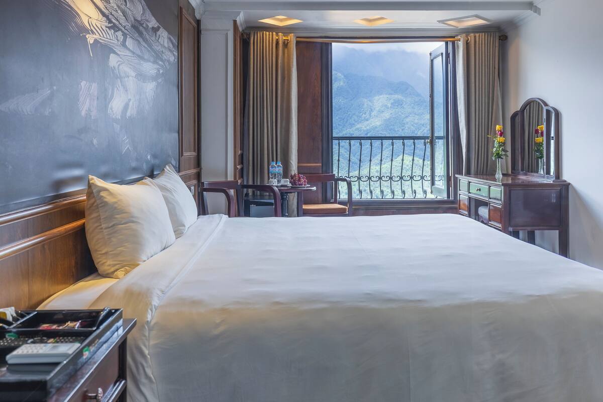 Habitación de hotel con una cama grande, un escritorio con una silla, una mesita con un florero y vistas a las montañas a través de la ventana.